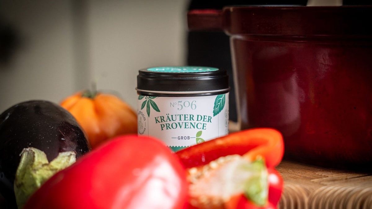 Kräuter der Provence im Glas, dazu frisches Gemüse und ein roter Topf auf dem Tisch.