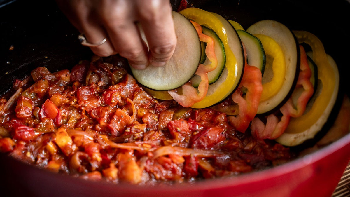Eine Hand legt geschnittenes Gemüse auf eine Tomaten-Zwiebel-Mischung in einem roten Kochtopf.