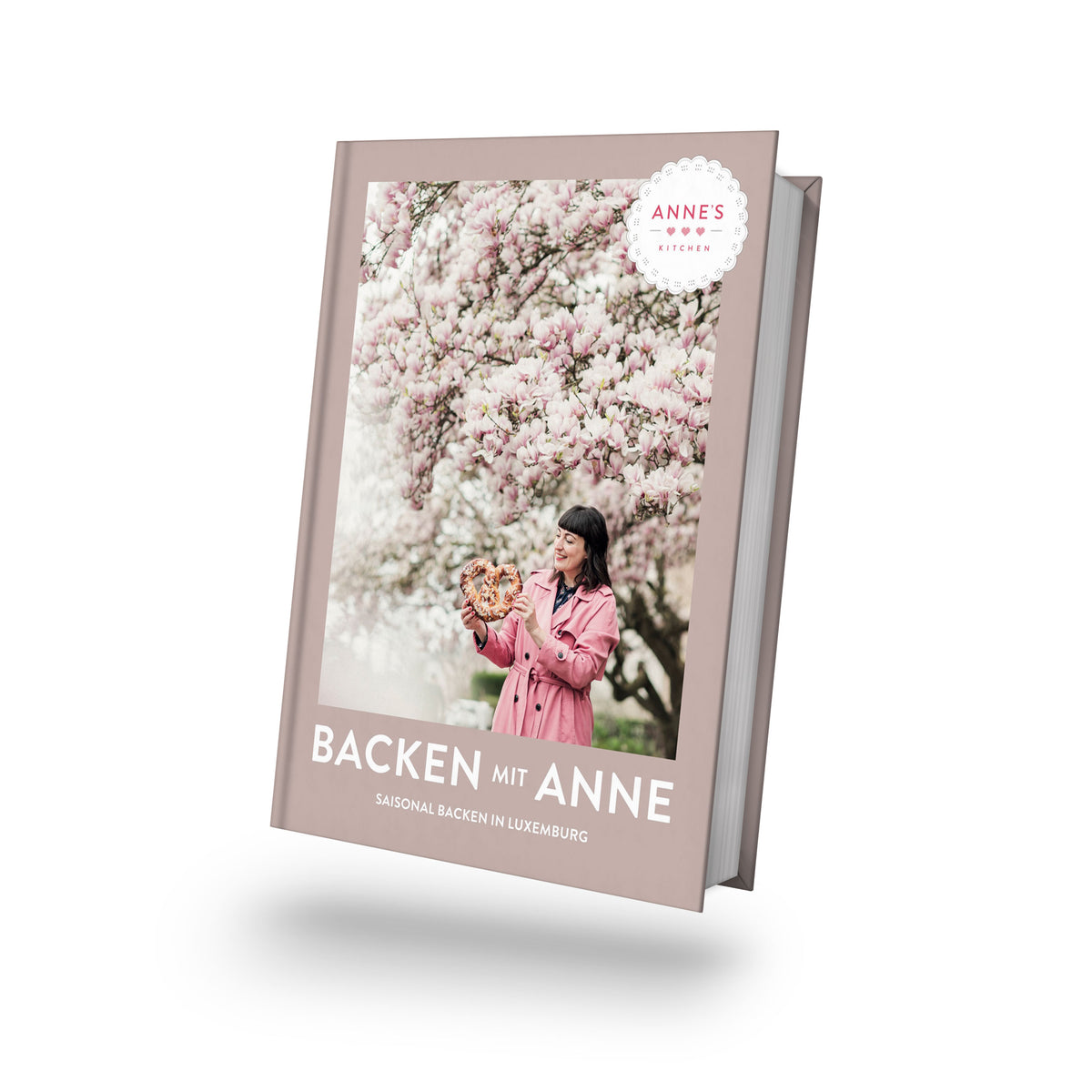 Frau im rosa Mantel hält saisonale Backwaren unter Blüten, Titel: Backen mit Anne.