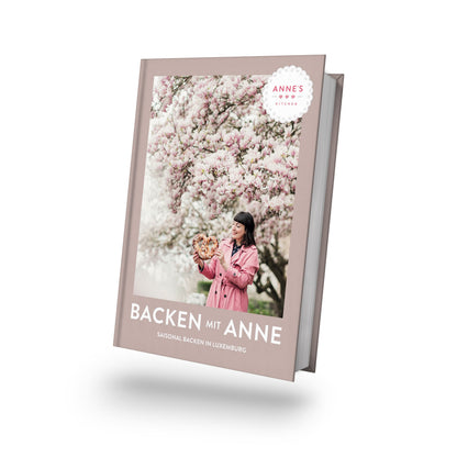 Frau im rosa Mantel hält saisonale Backwaren unter Blüten, Titel: Backen mit Anne.