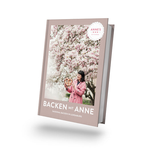 Frau im rosa Mantel hält saisonale Backwaren unter Blüten, Titel: Backen mit Anne.