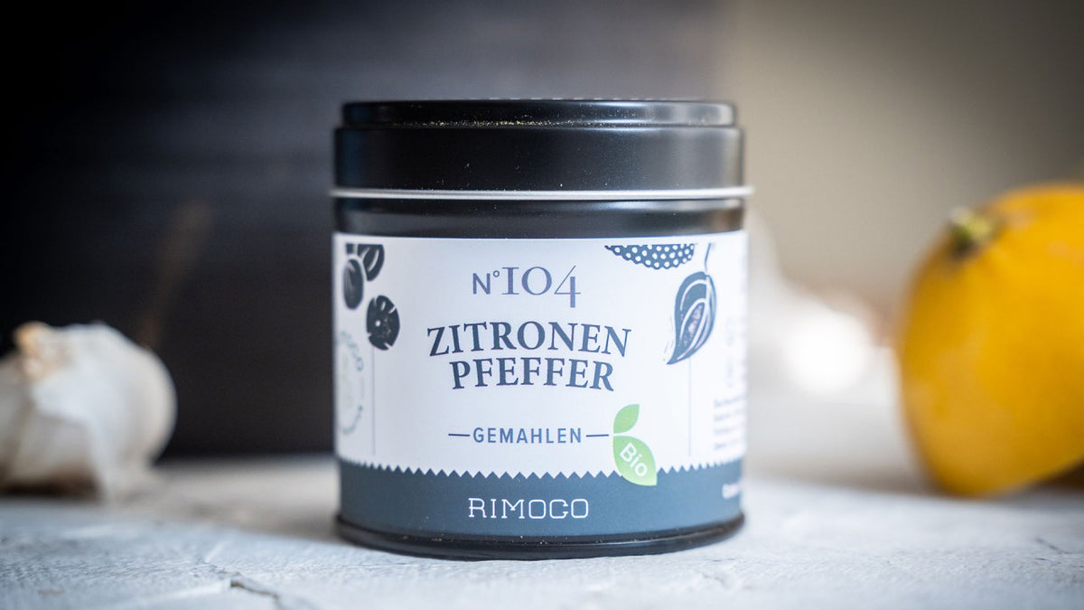 Eine Zitronen-Pfeffer-Dose steht neben einer frischen Zitrone und Knoblauch auf einer weißen Fläche.