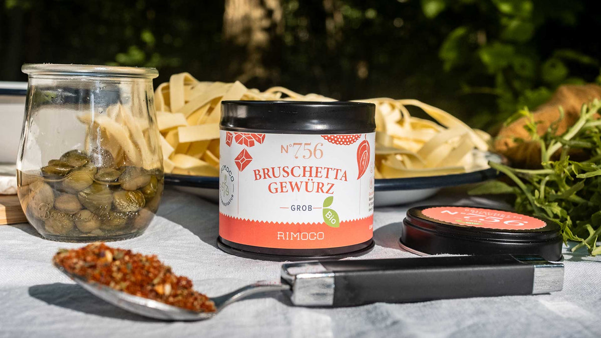 Ein Glas mit Kapern, Bruschetta-Gewürz, Pasta und ein Löffel mit Gewürzmischung auf einem Holztisch.