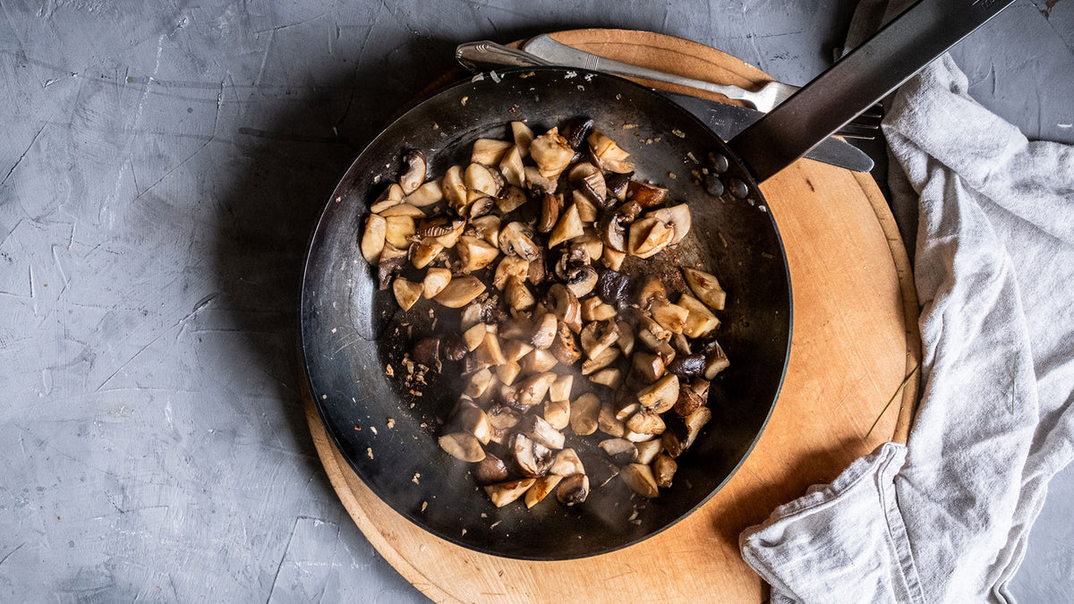 Gebratene Champignons in einer Pfanne auf einem Holzbrett, daneben ein hellgraues Stofftuch von oben.