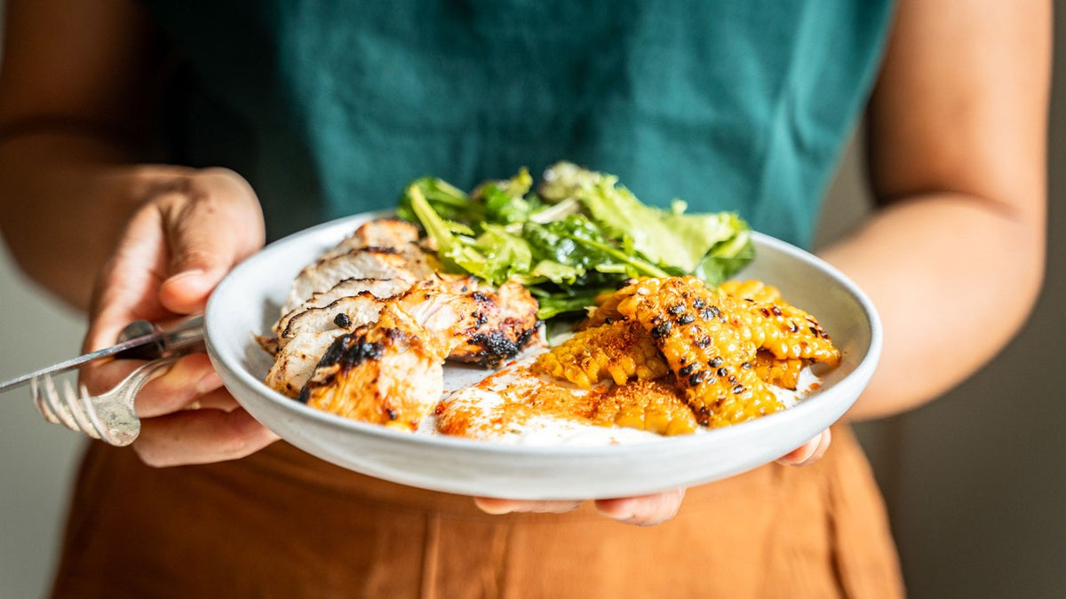 Gegrilltes Hähnchen, Mais und Salat auf einem weißen Teller, der von Händen gehalten wird.