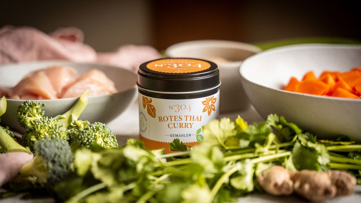 Ein Glas mit rotem Thai-Curry-Gewürz steht auf einem Holztisch zwischen frischem Gemüse und rohem Hähnchen.