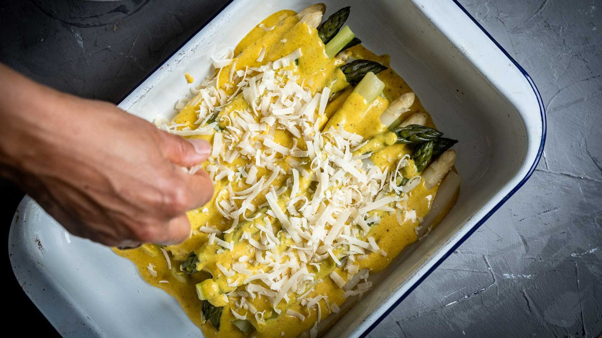 Eine Hand streut Käse über Spargel mit gelber Soße in einer weißen Auflaufform.
