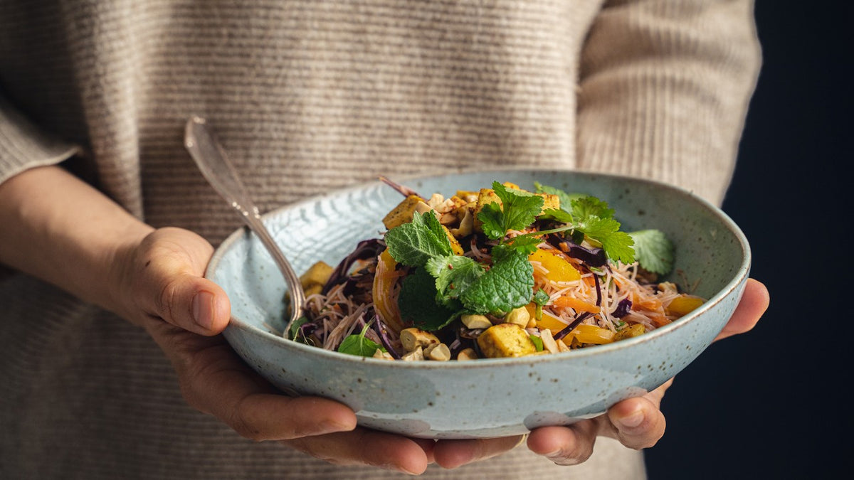 Eine Person hält einen bunten Nudelsalat mit frischen Kräutern, knackigen Nüssen und Gemüse in der Hand.