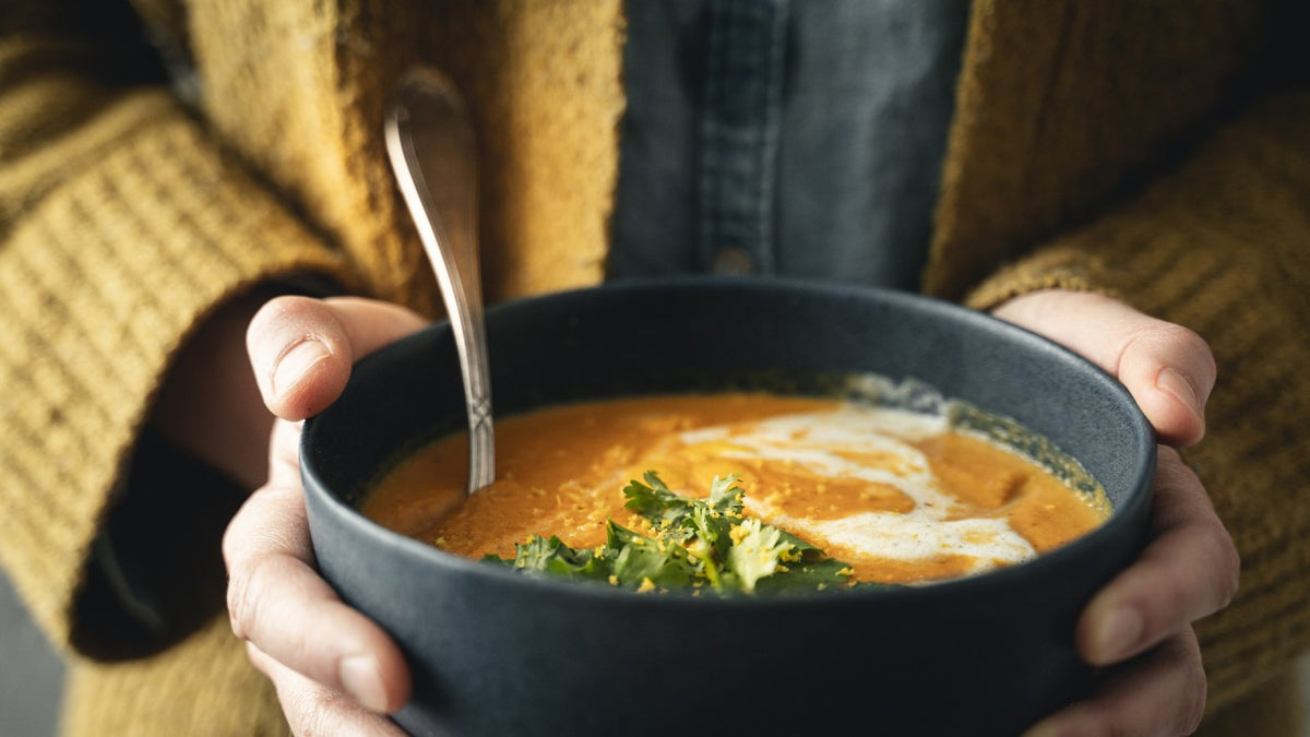 Eine Schüssel mit orangefarbener Suppe, darauf Kräuter und Sahne, von einer Person mit einem Löffel gehalten.