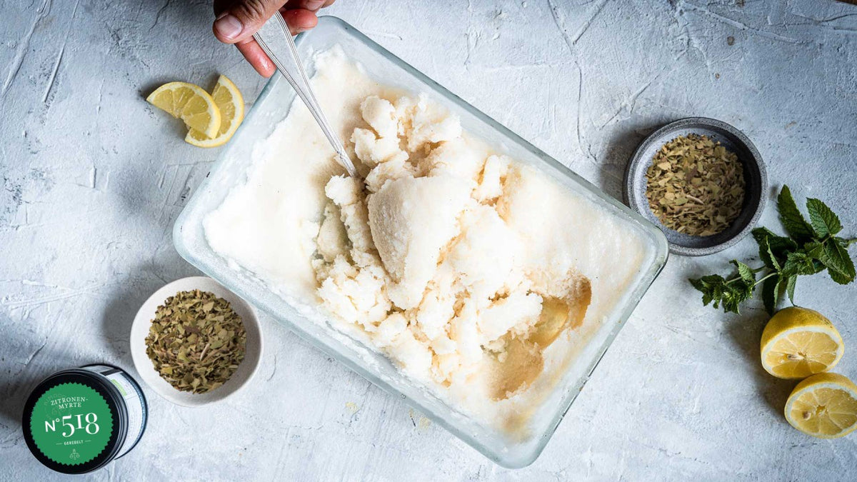 Eine Hand schöpft Granita aus einer Glasschale, daneben liegen Zitronen, Minze und Schalen mit Fenchelsamen.