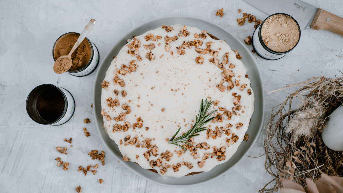 Runder Kuchen mit weißer Glasur und Nüssen, garniert mit Rosmarin auf einem grauen Teller neben Gewürzen.