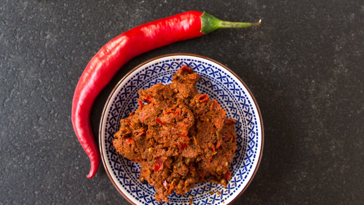 Eine rote Chilischote und eine gemusterte Schale mit roter Currypaste auf dunklem Hintergrund.