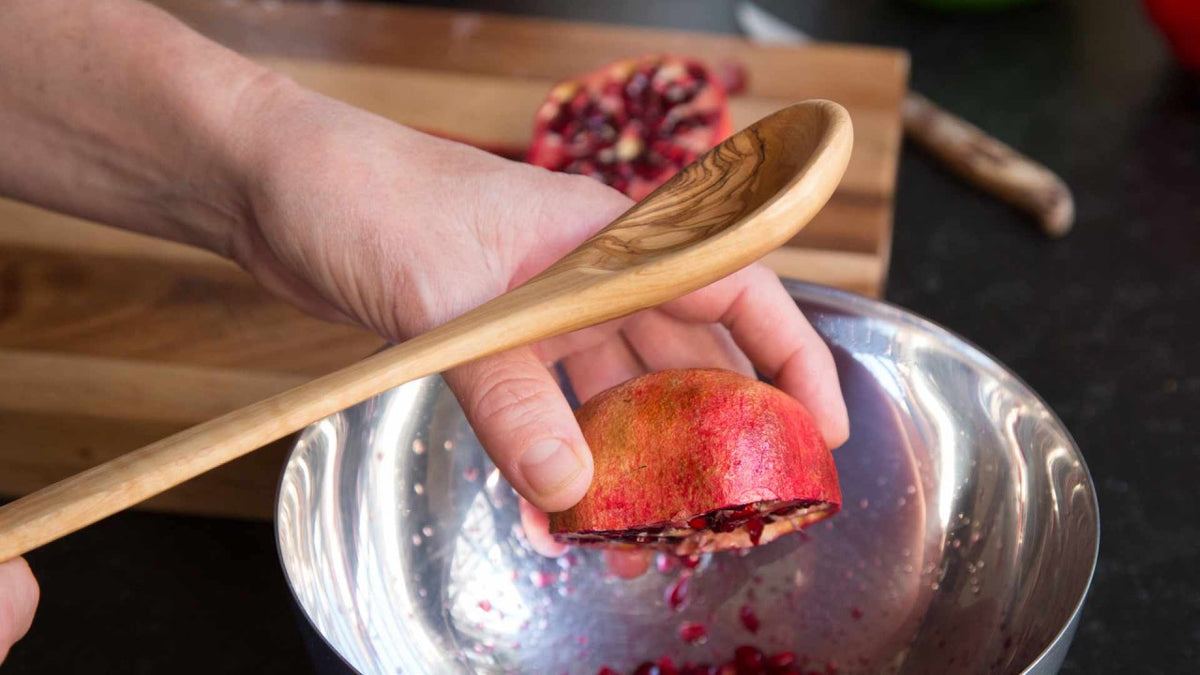 Eine Hand schlägt mit einem Holzlöffel auf eine Granatapfelhälfte über einer Metallschüssel.