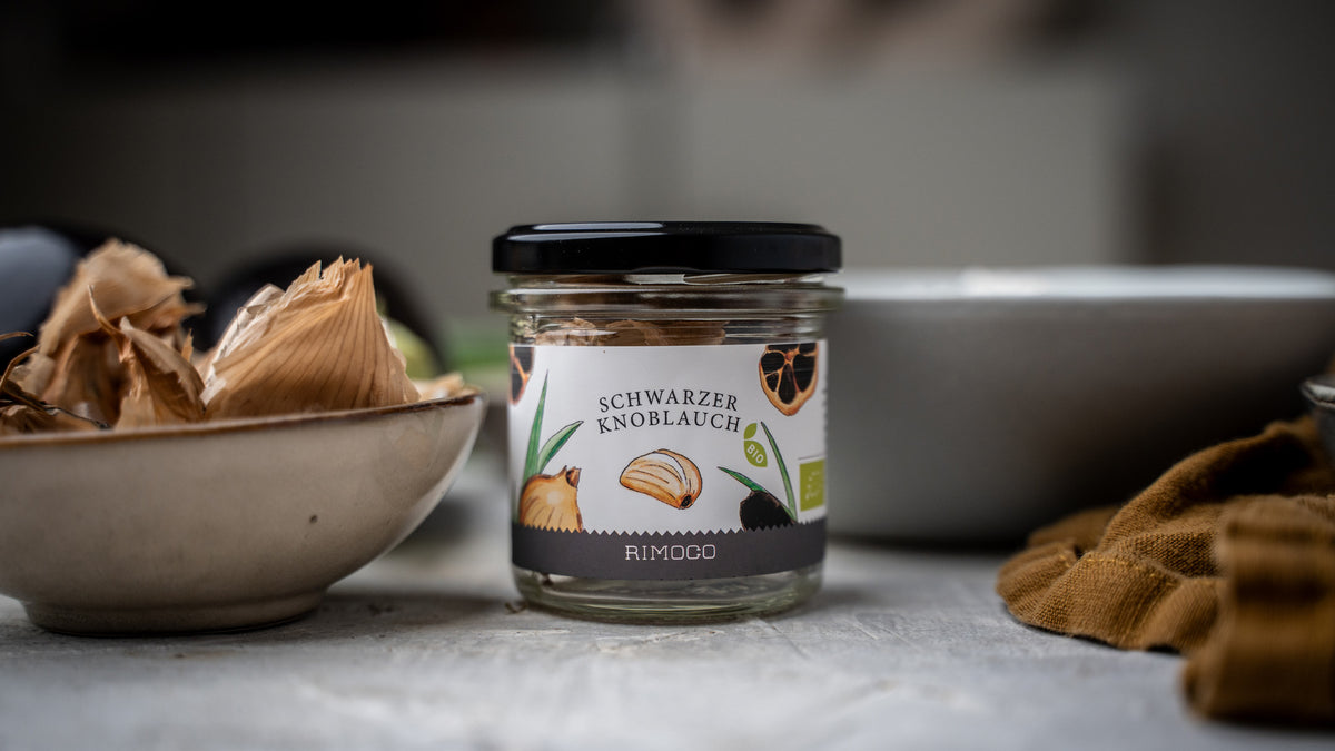 Bio Schwarze Knoblauchpaste im Glas steht neben Schalen und einem Tuch auf einer Oberfläche.