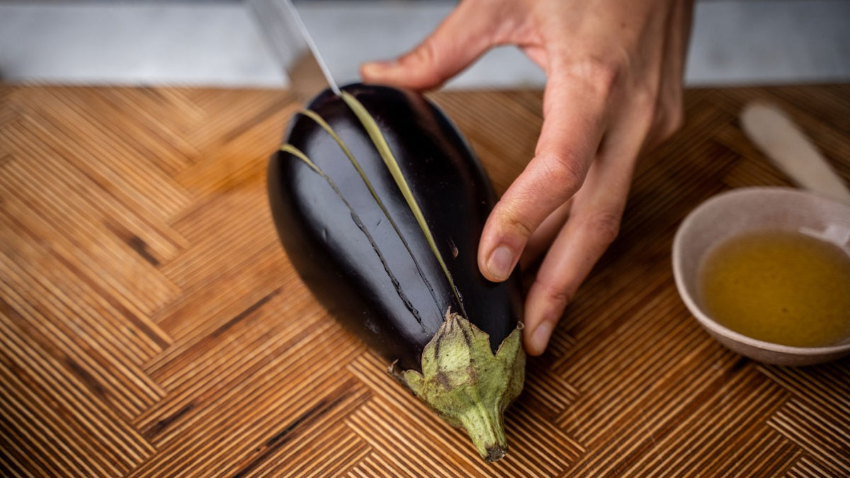Eine Hand schneidet eine Aubergine auf einem Holzbrett, daneben steht eine kleine Schale mit Olivenöl.