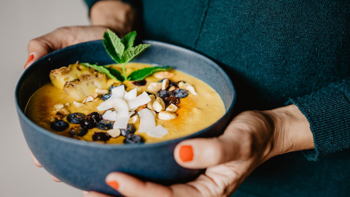 Eine Person hält eine bunte Smoothie-Bowl mit frischem Obst, Nüssen, Kokosflocken und Minze in der Hand.