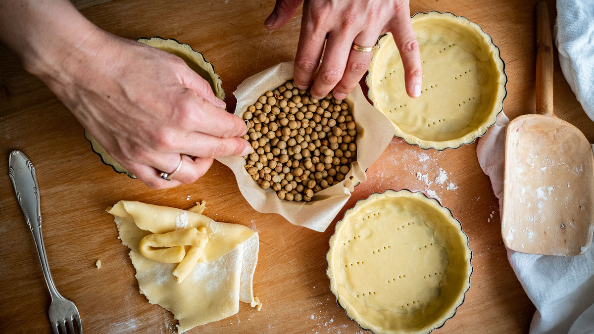 Auf einer rustikalen Holzfläche werden Tarteböden mit Backbohnen, Teig und Mehl vorbereitet.