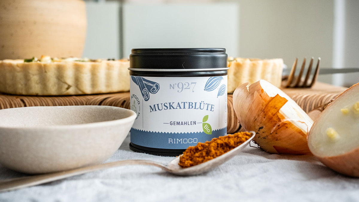 Eine Tarte, eine Zwiebel, ein Löffel Gewürz und eine Dose gemahlene Muskatblüte auf einem Holztisch.