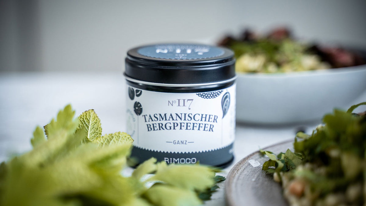 Eine Dose tasmanischer Bergpfeffer, dazu frische Kräuter und bunte Pfefferkörner im Hintergrund.