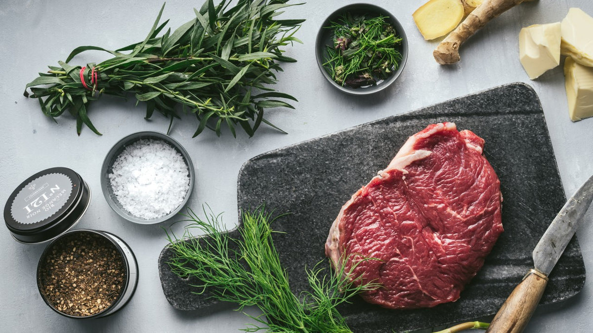 Rohes Steak auf Steinplatte mit frischen Kräutern, grobem Salz, Pfeffer, Butter und Kochmesser.