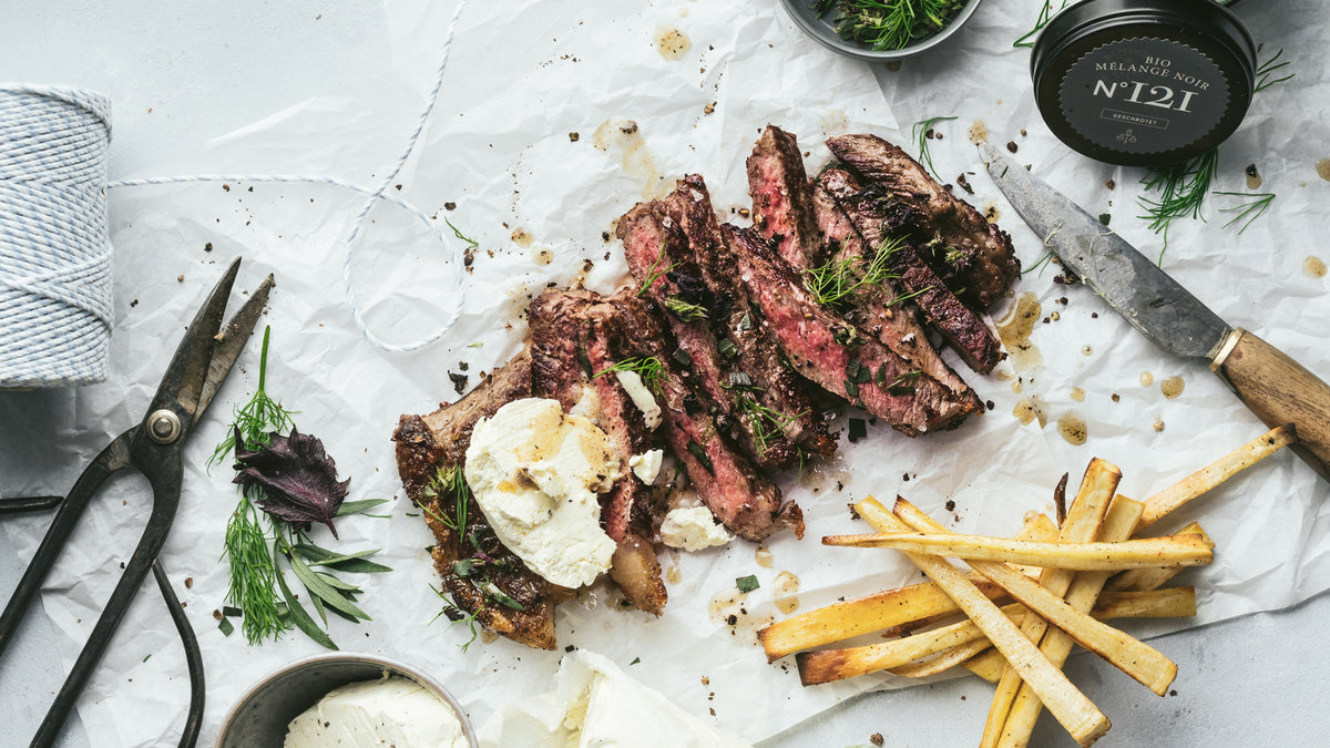 Geschnittenes Steak mit Kräutern, Käse und grobem Pfeffer, dazu Pommes auf Backpapier.