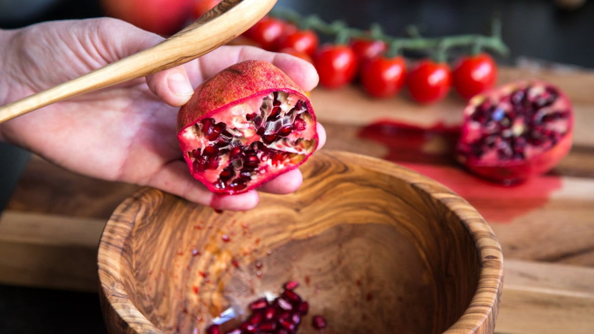 Eine Hand hält einen aufgeschnittenen Granatapfel über eine Holzschale, im Hintergrund sind Tomaten am Zweig zu sehen.
