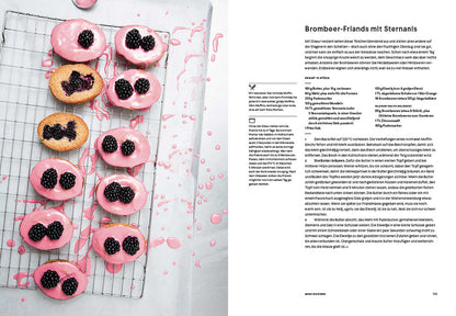 Mehrere ovale Gebäckstücke mit rosa Zuckerguss und Brombeeren auf einem Gitter neben einem Buch.