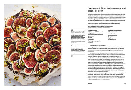 Eine Pavlova mit Feigen und Pistazien neben einer aufgeschlagenen Seite eines Rezeptbuchs.