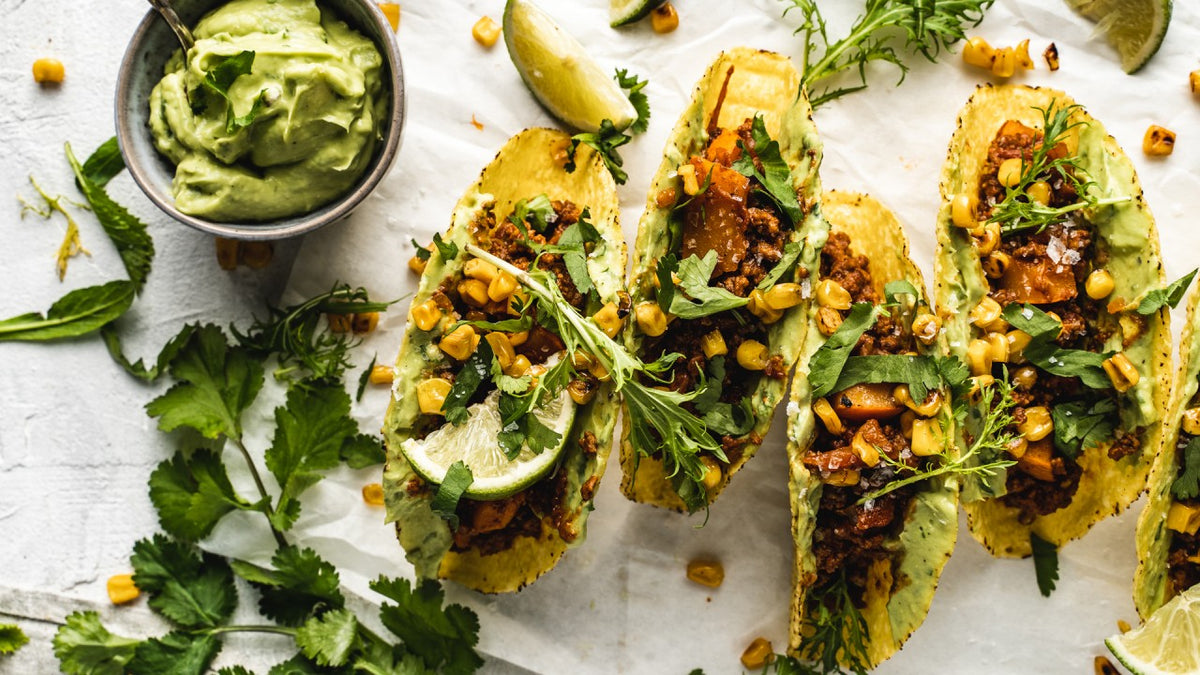 Drei Tacos mit gewürzter Füllung, frischem Mais, Gemüse und einer Schale cremigem Dip dazu.