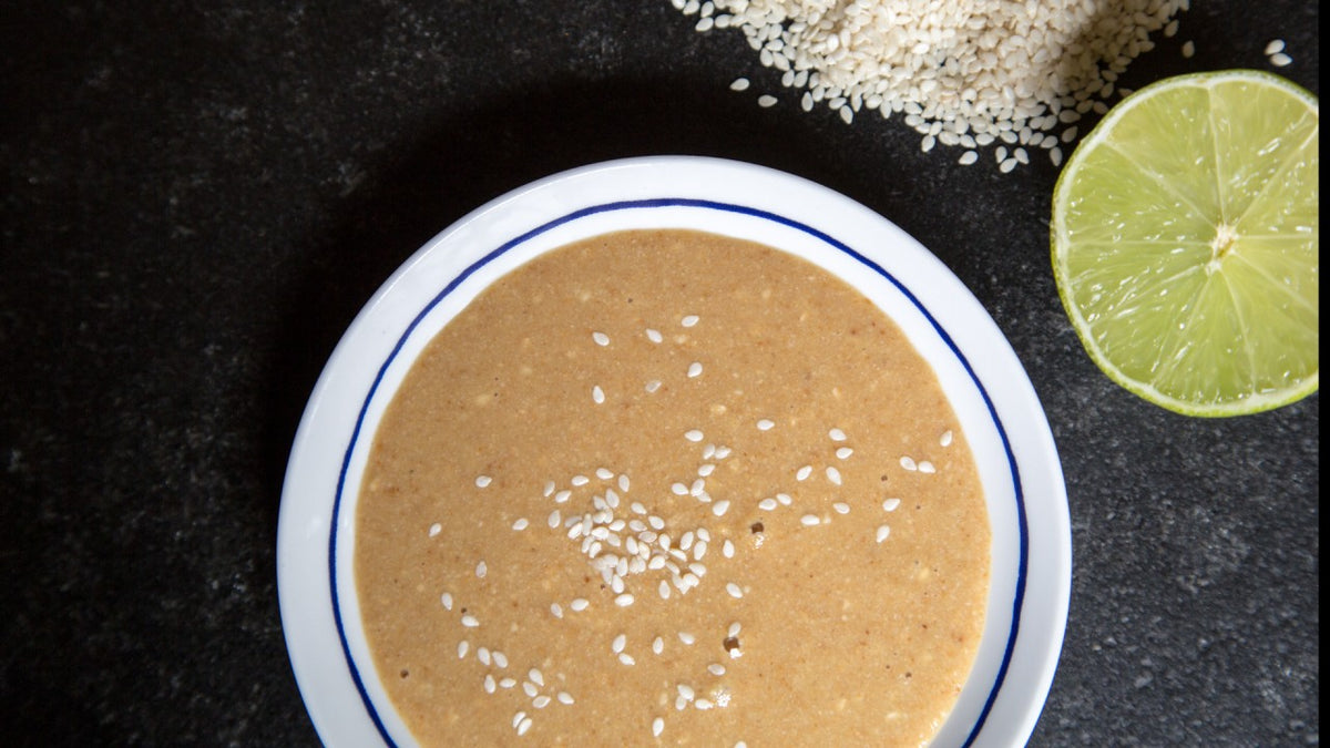 Eine Schale Tahini mit Sesamkörnern, daneben eine halbe Limette und weitere geröstete Sesamkörner.
