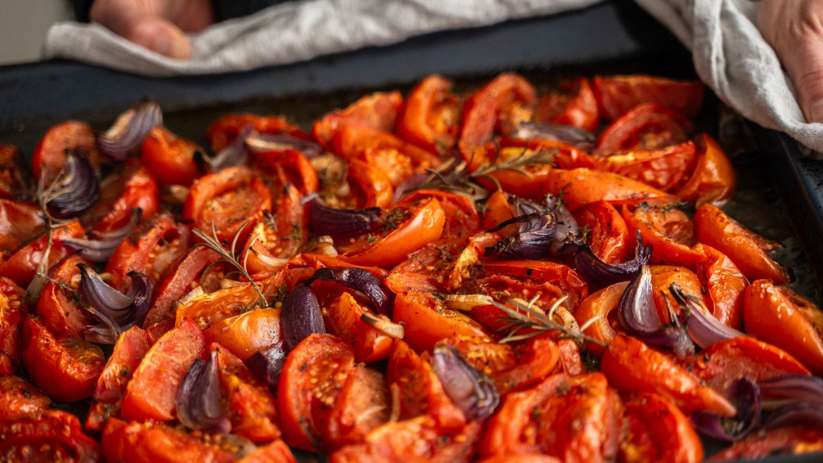 Geröstete Tomaten- und rote Zwiebelspalten mit Kräutern auf einem Backblech, gehalten von Händen, die ein Tuch umschließen.