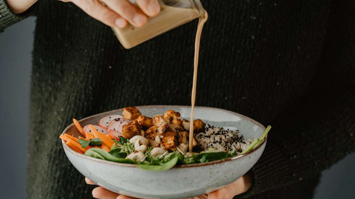 Eine Person gießt Dressing über einen Salat mit Tofu, Quinoa und frischen grünen Blättern.