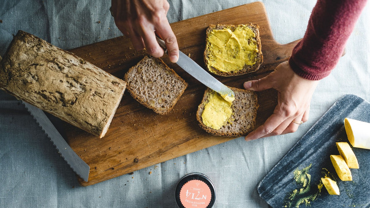 Eine Person bestreicht Vollkornbrotscheiben auf einem Holzbrett mit gelber Butter, im Hintergrund ist ein Brotlaib zu sehen.