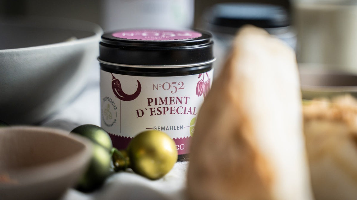 Auf einem Tisch steht ein Glas mit dem Etikett „Piment d’Especial“, daneben liegen unscharf abgebildete Gewürze und Dekoartikel.