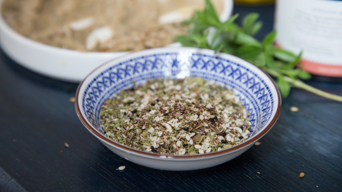 Auf einem dunklen Tisch steht eine Schale mit gemahlenem Za’atar-Gewürz, im Hintergrund frische Kräuter.