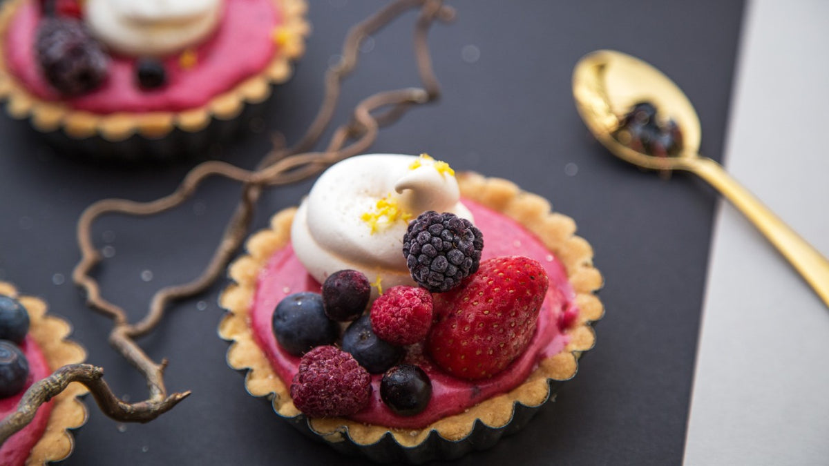 Eine Beerentarte mit Sahne und gefrorenen Beeren liegt neben einem goldenen Löffel auf einer Fläche.