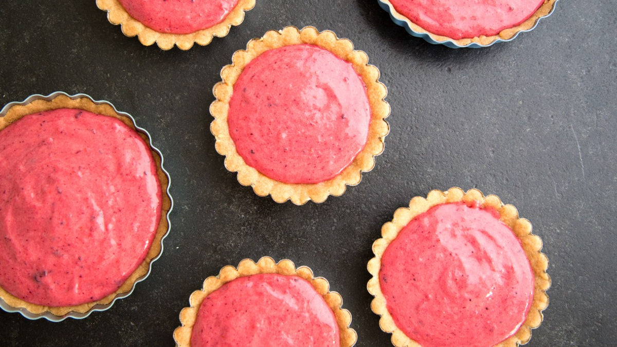 Sechs Mini-Törtchen mit leuchtend rosa Beerencreme, dekorativ auf einem dunklen Untergrund angeordnet.